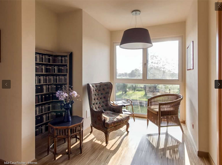 Cozy reading nook with a vintage armchair, wicker chair, and a small table with flowers, beside a window overlooking greenery.