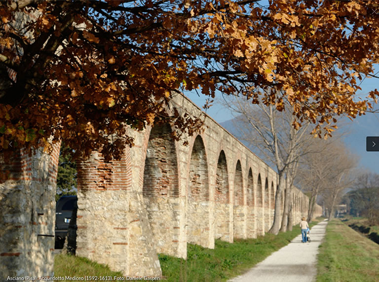 Walk-path beside a historic brick aqueduct, framed by autumn leaves and gentle sunlight.