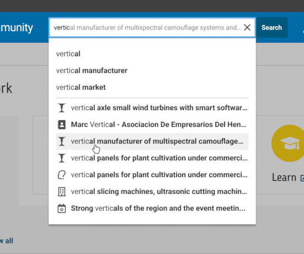 Search bar interface displaying autocomplete suggestions for the query "vertical manufacturer of multispectral camouflage systems and...". The dropdown list includes related search terms such as "vertical manufacturer", "vertical market", and "vertical axle small wind turbines with smart software". A cursor hovers over the suggestion "vertical manufacturer of multispectral camouflage...". The search is conducted on a website with a blue navigation bar and a partially visible "Learn" icon featuring a graduation cap. This image illustrates user search behavior, keyword input, and predictive text features on a professional or technical platform.